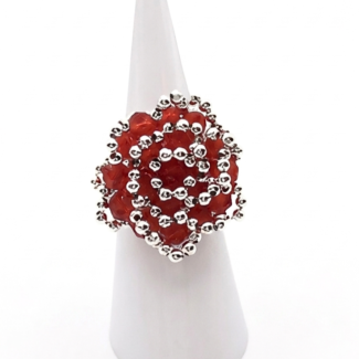 Handmade oversized cocktail ring featuring red faceted crystals and silver beads woven in a spiral rose pattern, worn on a finger of a mannequin hand against a white background