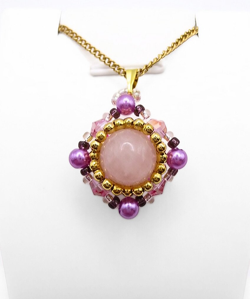 Handmade rose quartz pendant featuring a large smooth round centre stone surrounded by pink crystals, vibrant purple pearls, and gold seed beads in a diamond-shaped silhouette, with a gold-tone bail, shown against a white display stand.