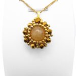 Handmade reversible tourmaline pendant front view featuring a smooth round peach tourmaline centre stone encircled by metallic gold-plated faceted crystals and gold bead accents in a sunburst floral design, with a gold-tone bail, shown on a white display stand.