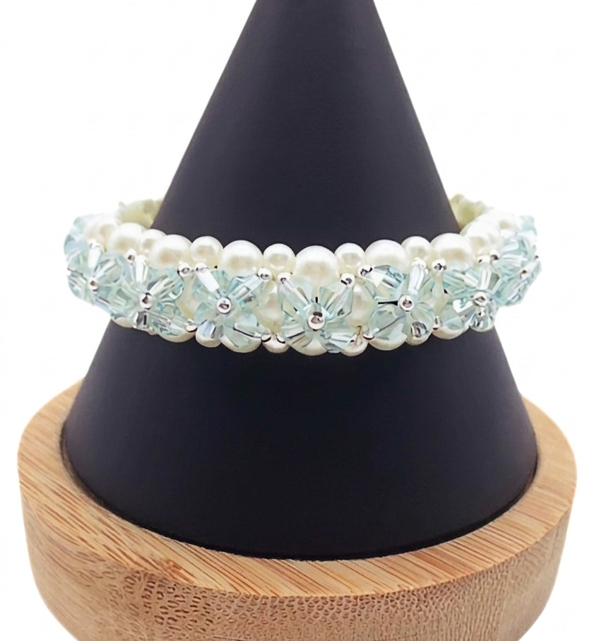 A close-up photograph of a hand-woven multi-strand beaded bracelet on a black display hand against a plain white background. The bracelet is made with white faux pearls and faceted light blue crystal beads arranged in a floral weave with silver bead accents.