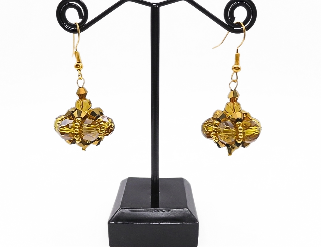 Handmade cluster drop earrings featuring large translucent golden crystals and smaller metallic gold crystals woven into a pendulum cluster shape, with gold bead accents and gold-tone ear wires, displayed on a black metal earring stand.