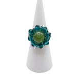 Handmade beaded cocktail ring featuring an olive green tourmaline centre stone surrounded by teal blue and emerald green crystal beads, displayed on a white cone ring stand