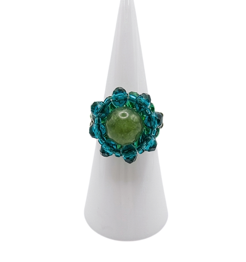 green-tourmaline-ring-display Handmade beaded cocktail ring featuring an olive green tourmaline centre stone surrounded by teal blue and emerald green crystal beads, displayed on a white cone ring stand