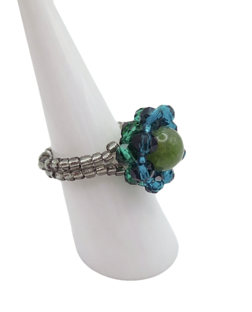 green-tourmaline-ring-side-view Side view of handmade beaded ring showing silver seed bead band construction and layered teal blue and green crystal halo surrounding an olive green tourmaline centre stone