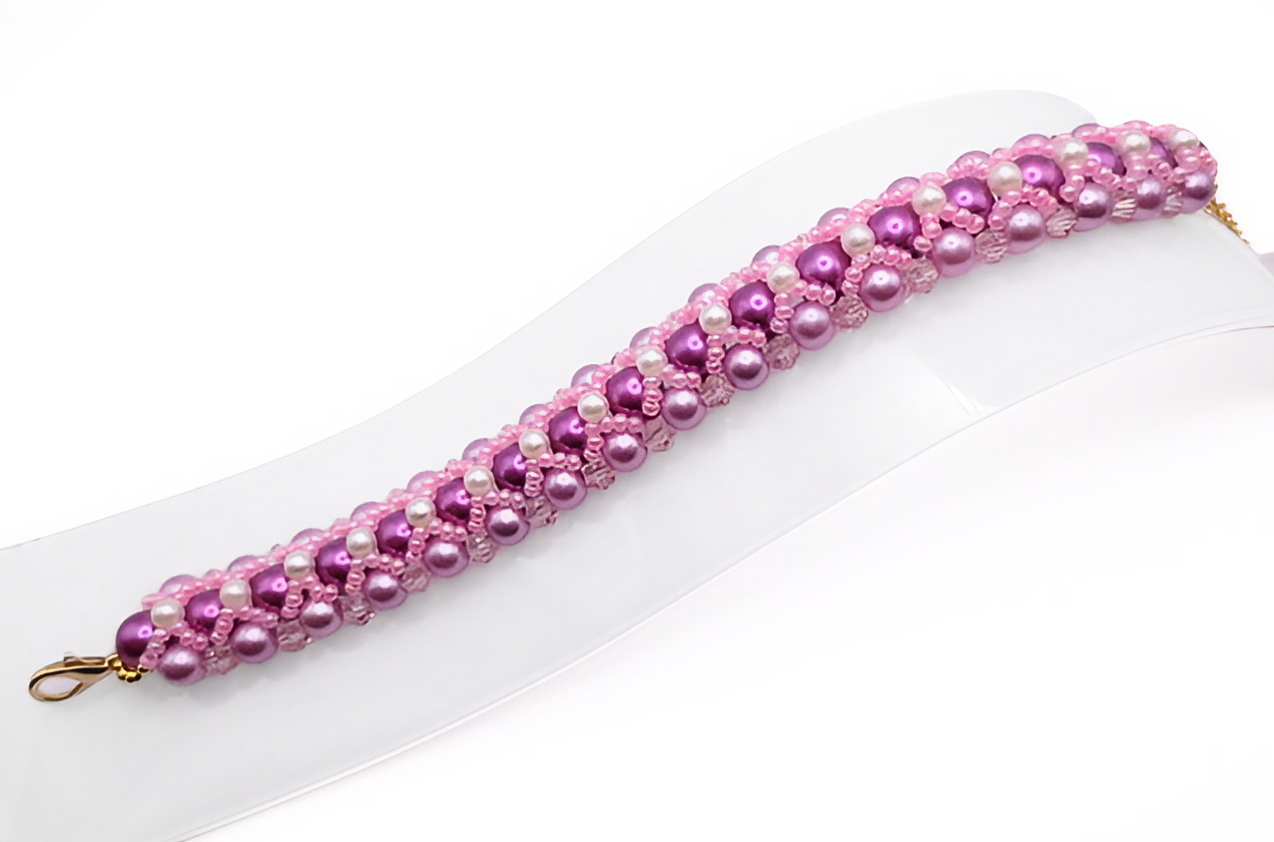 Wide bracelet featuring deep purple and lavender pearls woven together with small white pearl accents, bright pink beads and light pink crystal detailing in a repeating diagonal pattern, with a gold-tone lobster clasp, displayed flat on a white jewellery stand against a white background.