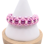 Handmade wide purple and pink pearl bracelet featuring deep purple and lavender pearls, small white pearl accents and bright pink beads and light pink crystal detailing in a full rounded woven design, displayed on a white cone bracelet stand with a wooden base against a white background.