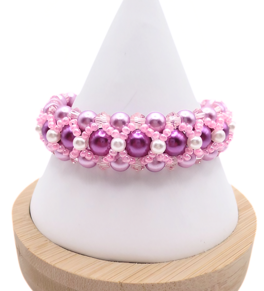 Handmade wide purple and pink pearl bracelet featuring deep purple and lavender pearls, small white pearl accents and bright pink beads and light pink crystal detailing in a full rounded woven design, displayed on a white cone bracelet stand with a wooden base against a white background.