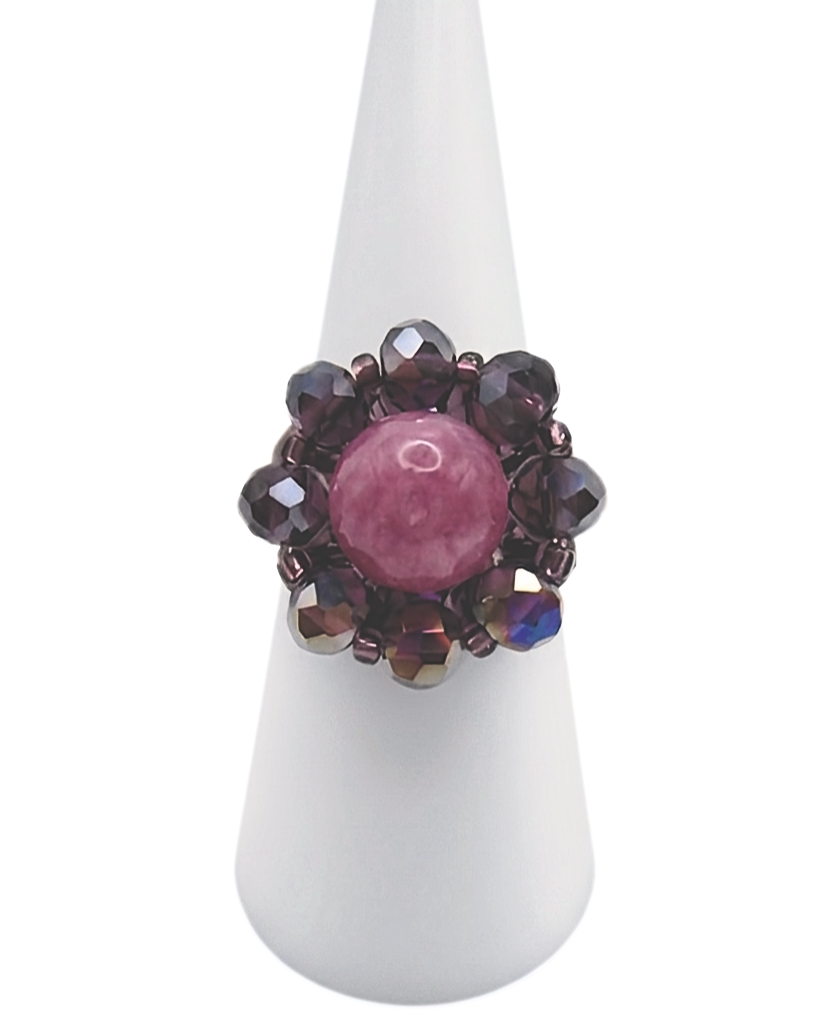 rose-tourmaline-ring-display Handmade beaded cocktail ring featuring a round rose pink tourmaline centre stone surrounded by faceted purple AB crystal beads in a floral halo design, displayed on a white cone ring stand