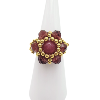 Handmade tourmaline and crystal cocktail ring with gold bead halo, featuring a round rose-pink tourmaline centre stone surrounded by faceted burgundy crystals and gold seed beads, displayed on white cone ring stand