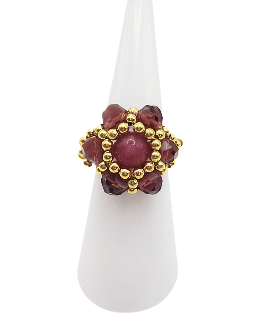 tourmaline-ring-display Handmade tourmaline and crystal cocktail ring with gold bead halo, featuring a round rose-pink tourmaline centre stone surrounded by faceted burgundy crystals and gold seed beads, displayed on white cone ring stand