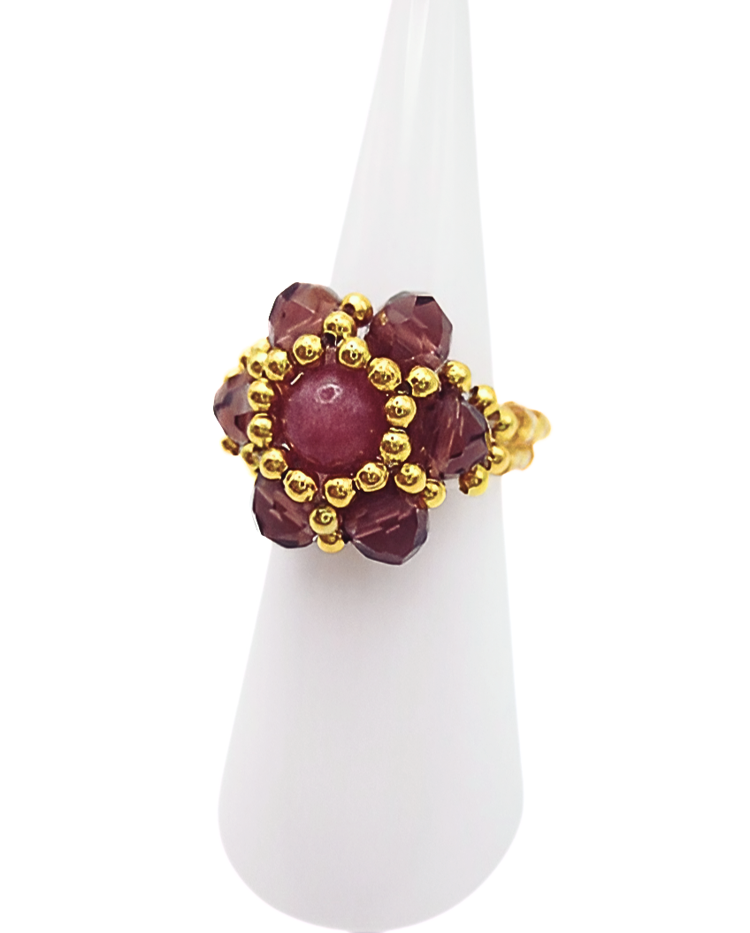 tourmaline-ring-side-view Angled side view of handmade pink tourmaline beaded ring with burgundy crystal petals and gold bead halo on gold seed bead band, displayed on white ceramic ring cone
