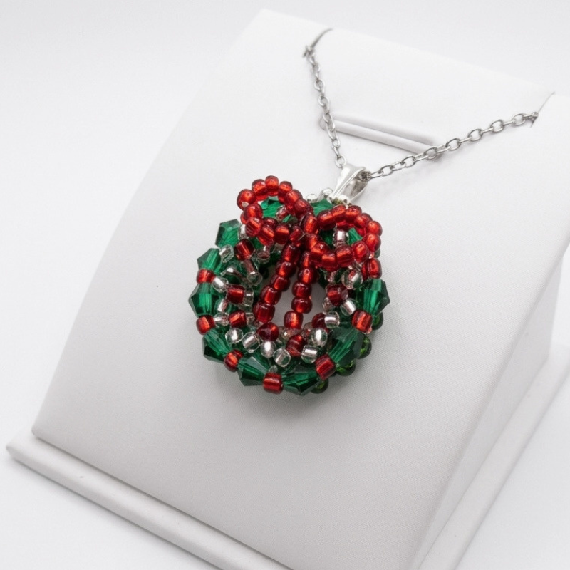 christmas-wreath-pendant-angled-view Angled view of a handcrafted green crystal holiday wreath pendant with red beaded bow, displayed on a white jewellery stand