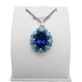 Handmade sapphire blue oval crystal pendant with a large faceted deep blue centre stone surrounded by aquamarine and light blue crystals with silver bead accents and a silver-tone bail, photographed on a display stand.
