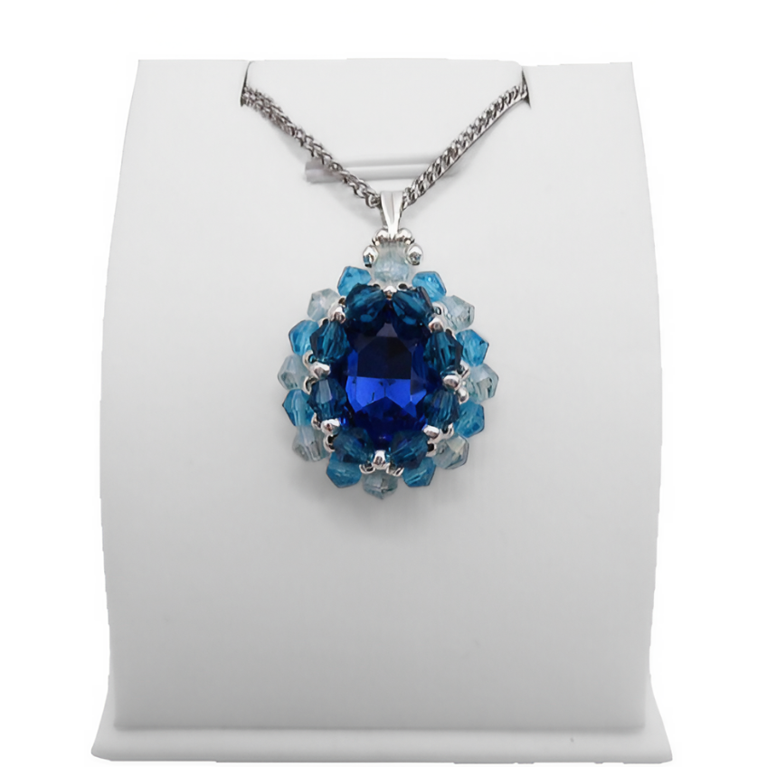 Handmade sapphire blue oval crystal pendant with a large faceted deep blue centre stone surrounded by aquamarine and light blue crystals with silver bead accents and a silver-tone bail, photographed on a display stand.