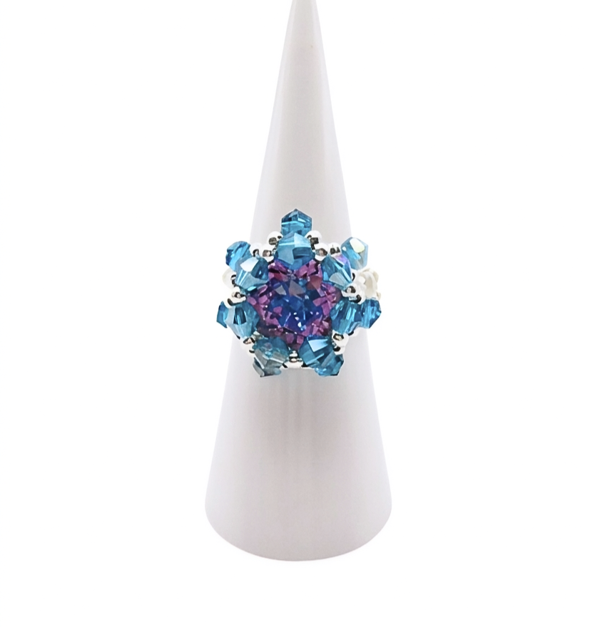 Handmade beaded cocktail ring featuring a purple and blue crystal centre surrounded by faceted ocean blue AB crystals and silver beads in a floral starburst design, displayed on a white cone ring stand