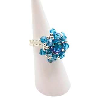 Side view of handmade ocean blue and purple crystal starburst cocktail ring on white cone ring stand, showing double-row white and silver seed bead band construction and layered crystal detail
