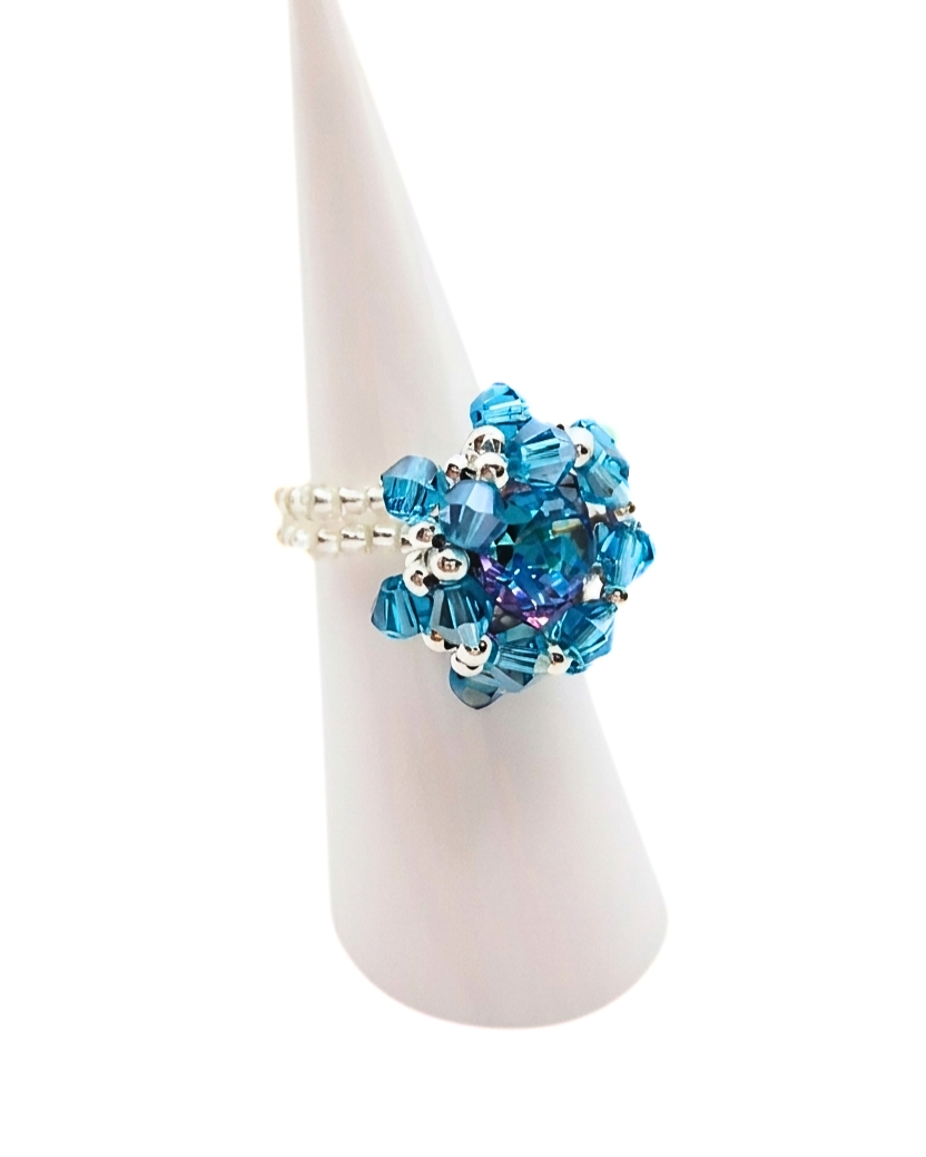 Side view of handmade ocean blue and purple crystal starburst cocktail ring on white cone ring stand, showing double-row white and silver seed bead band construction and layered crystal detail