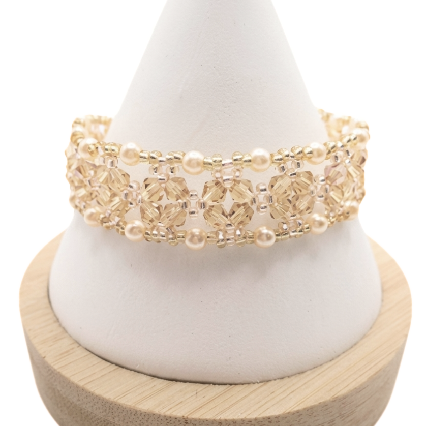 Handmade crystal bracelet featuring champagne faceted crystals and lustrous champagne pearls woven together with gold bead accents in an open lattice diamond pattern, displayed on a white cone bracelet stand with a wooden base against a white background.
