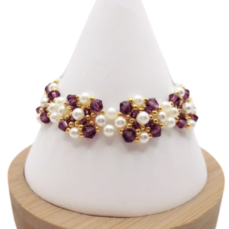 Handmade bracelet featuring deep purple faceted crystals and lustrous white pearls woven together in a repeating pattern with gold bead accents throughout, displayed on a white cone bracelet stand with a wooden base against a white background.