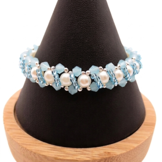 Handmade bracelet featuring pastel sky blue faceted crystals and large lustrous white pearls woven together in a repeating cluster pattern with silver-tone seed bead accents, displayed on a black cone bracelet stand with a wooden base against a white background.