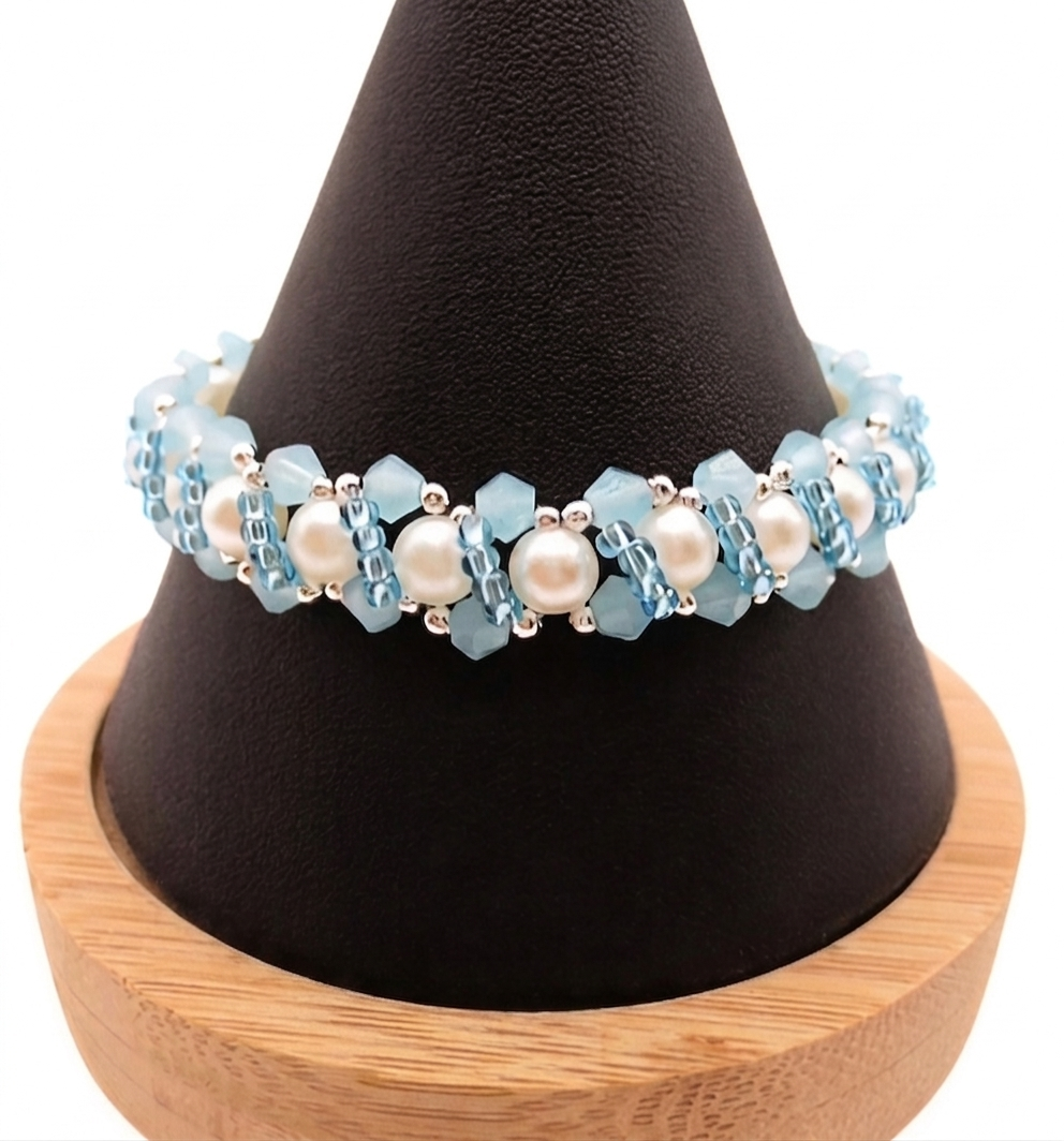 Handmade bracelet featuring pastel sky blue faceted crystals and large lustrous white pearls woven together in a repeating cluster pattern with silver-tone seed bead accents, displayed on a black cone bracelet stand with a wooden base against a white background.