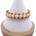 Handmade bracelet featuring purple faceted crystals and large lustrous white pearls woven together in a repeating cluster pattern with gold seed bead accents, displayed on a white cone bracelet stand with a wooden base against a white background.