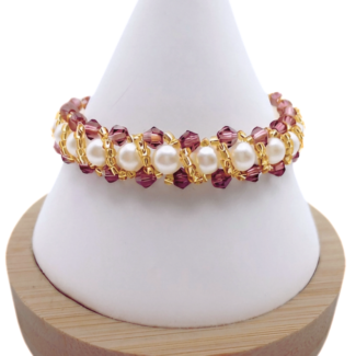 Handmade bracelet featuring purple faceted crystals and large lustrous white pearls woven together in a repeating cluster pattern with gold seed bead accents, displayed on a white cone bracelet stand with a wooden base against a white background.