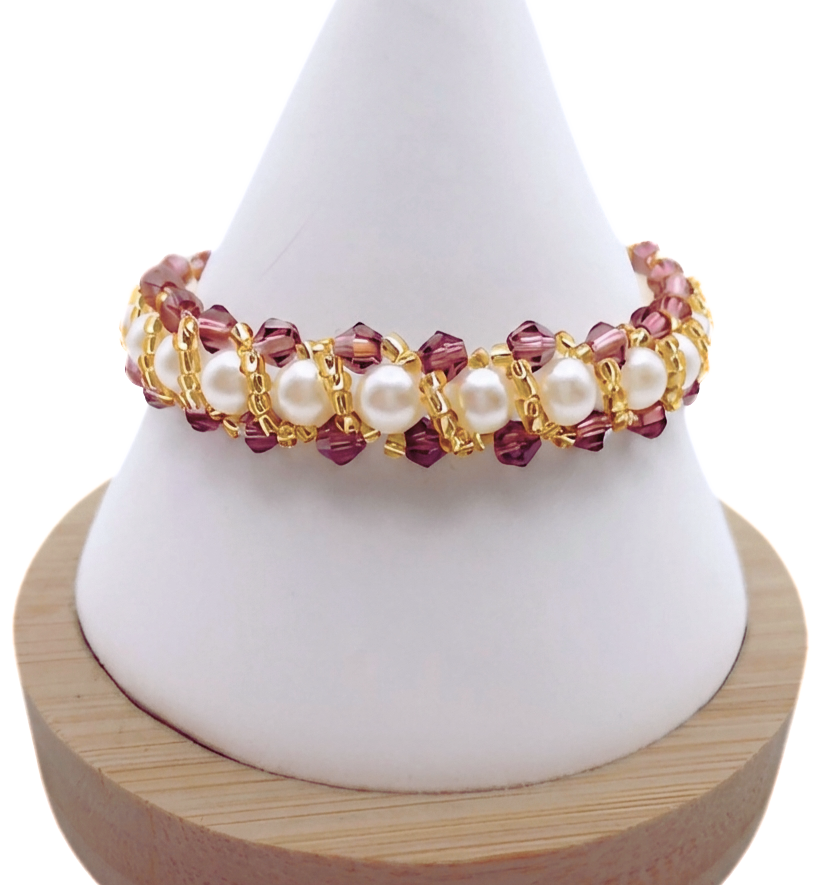 Handmade bracelet featuring purple faceted crystals and large lustrous white pearls woven together in a repeating cluster pattern with gold seed bead accents, displayed on a white cone bracelet stand with a wooden base against a white background.