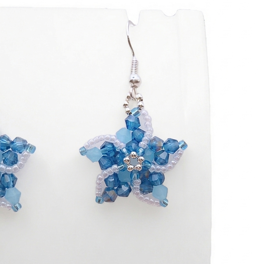 Close-up of a handmade crystal starfish earring displayed on a white earring stand, showcasing the intricate beadwork of pastel blue and iridescent teal AB-coated crystals with a silver centre detail.