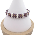 Handmade bracelet featuring violet faceted crystals arranged in a repeating four-petal flower pattern with white pearl centres and borders, and gold seed bead accents, displayed on a white cone bracelet stand with a wooden base against a white background.