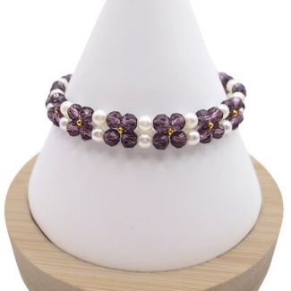 Handmade bracelet featuring violet faceted crystals arranged in a repeating four-petal flower pattern with white pearl centres and borders, and gold seed bead accents, displayed on a white cone bracelet stand with a wooden base against a white background.