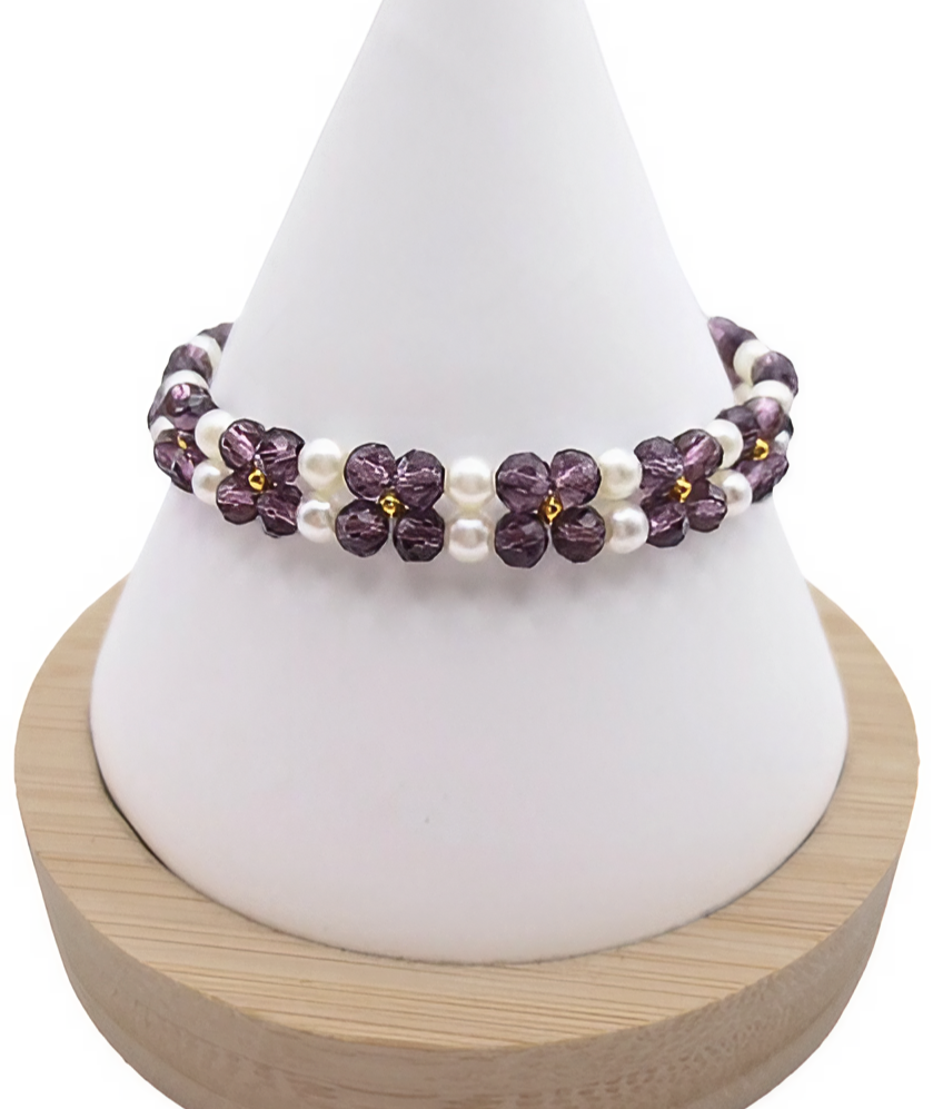 Handmade bracelet featuring violet faceted crystals arranged in a repeating four-petal flower pattern with white pearl centres and borders, and gold seed bead accents, displayed on a white cone bracelet stand with a wooden base against a white background.