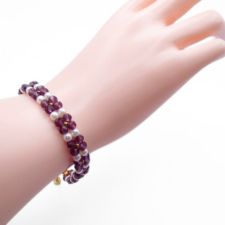 Handmade violet crystal and white pearl floral bracelet featuring repeating four-petal crystal flower motifs with pearl centres and gold seed bead accents, with a gold-tone lobster clasp, shown worn on a mannequin wrist and hand against a white background.