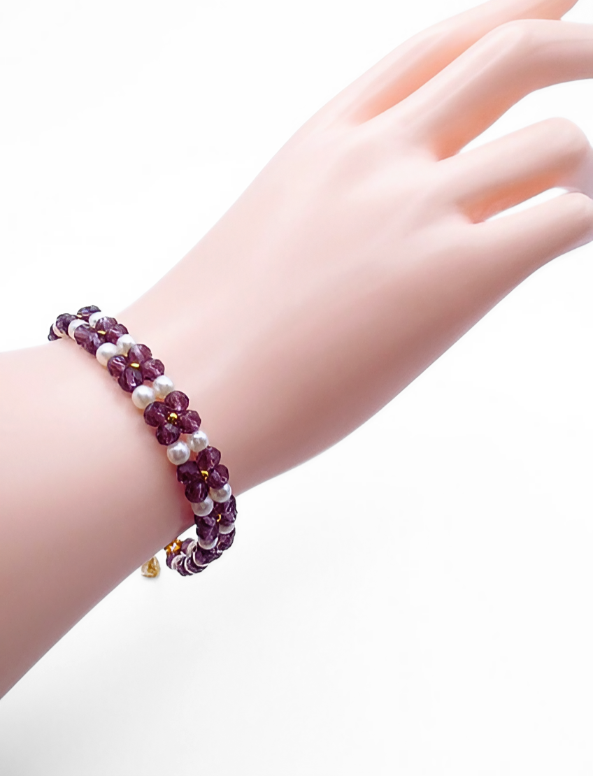 Handmade violet crystal and white pearl floral bracelet featuring repeating four-petal crystal flower motifs with pearl centres and gold seed bead accents, with a gold-tone lobster clasp, shown worn on a mannequin wrist and hand against a white background.