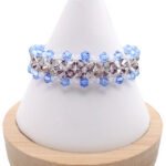 Handmade wide bracelet featuring alternating light blue and metallic grey faceted crystals woven together with clear seed bead accents in a repeating diamond lattice pattern, displayed on a white cone bracelet stand with a wooden base against a white background.