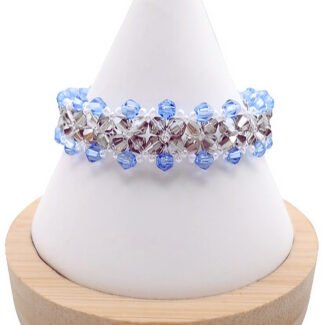Handmade wide bracelet featuring alternating light blue and metallic grey faceted crystals woven together with clear seed bead accents in a repeating diamond lattice pattern, displayed on a white cone bracelet stand with a wooden base against a white background.