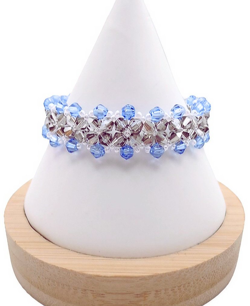 Handmade wide bracelet featuring alternating light blue and metallic grey faceted crystals woven together with clear seed bead accents in a repeating diamond lattice pattern, displayed on a white cone bracelet stand with a wooden base against a white background.