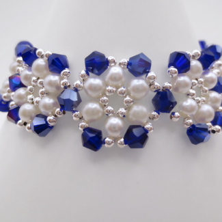 Close-up view of the handmade wide bracelet featuring deep metallic-blue AB reflective faceted crystals and lustrous white pearls woven together in a repeating floral-cluster pattern with silver-tone bead accents, shown against a white cone bracelet stand.