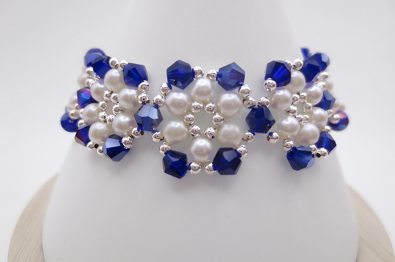 Close-up view of the handmade wide bracelet featuring deep metallic-blue AB reflective faceted crystals and lustrous white pearls woven together in a repeating floral-cluster pattern with silver-tone bead accents, shown against a white cone bracelet stand.