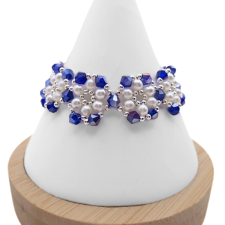 Handmade wide deep metallic blue AB crystal and white pearl bracelet featuring reflective blue faceted crystals and lustrous white pearls woven in a repeating floral cluster pattern with silver-tone bead accents, displayed on a white cone bracelet stand with a wooden base against a white background.
