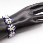 Handmade wide bracelet featuring deep metallic blue AB reflective faceted crystals and lustrous white pearls woven together in a repeating floral cluster pattern with silver-tone bead accents, shown worn on a black mannequin hand and wrist against a white background.