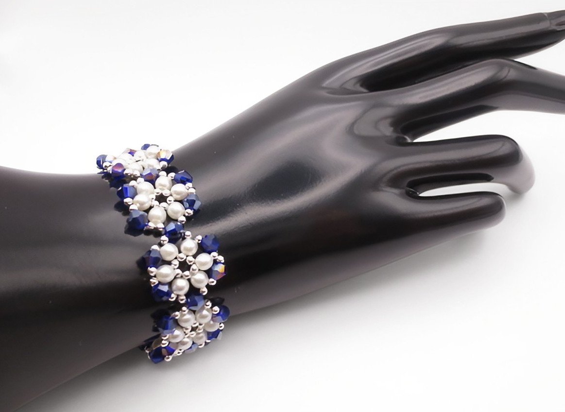 Handmade wide bracelet featuring deep metallic blue AB reflective faceted crystals and lustrous white pearls woven together in a repeating floral cluster pattern with silver-tone bead accents, shown worn on a black mannequin hand and wrist against a white background.