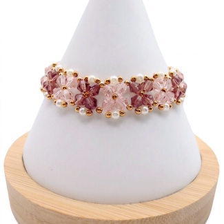 Handmade bracelet featuring light pink and purple faceted crystals arranged in a repeating four-petal flower pattern with small white pearl accents and gold seed bead detailing throughout, displayed on a white cone bracelet stand with a wooden base against a white background.