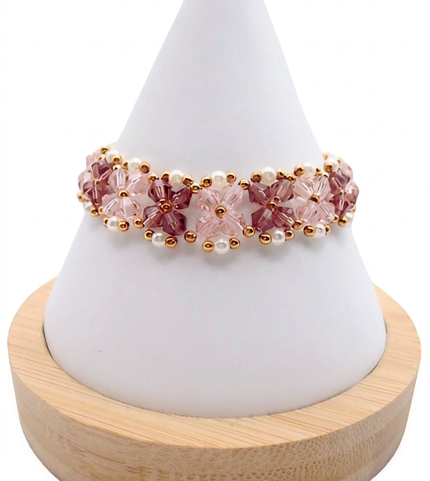 Handmade bracelet featuring light pink and purple faceted crystals arranged in a repeating four-petal flower pattern with small white pearl accents and gold seed bead detailing throughout, displayed on a white cone bracelet stand with a wooden base against a white background.