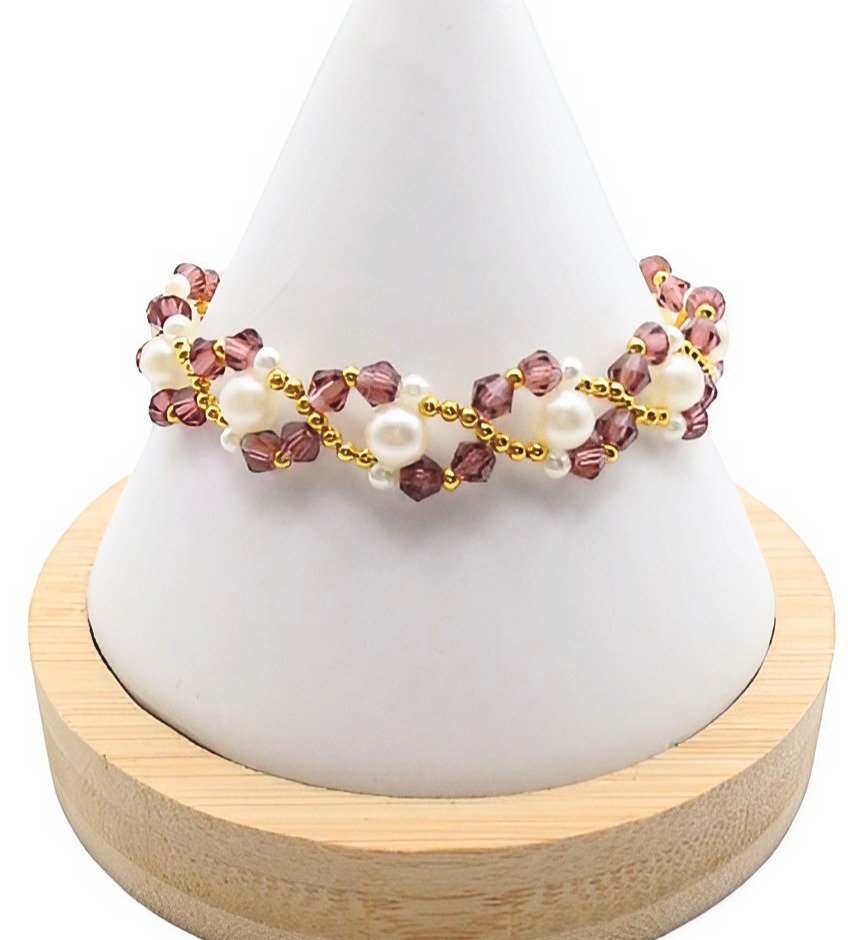 Handmade bracelet featuring purple faceted crystals and lustrous white pearls woven together in a zigzag pattern with gold seed bead accents throughout, displayed on a white cone bracelet stand with a wooden base against a white background.