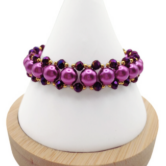 Handmade wide bracelet featuring large lustrous purple pearls and deep metallic purple faceted crystals woven together with gold seed bead accents in a full rounded cluster design, displayed on a white cone bracelet stand with a wooden base against a white background.