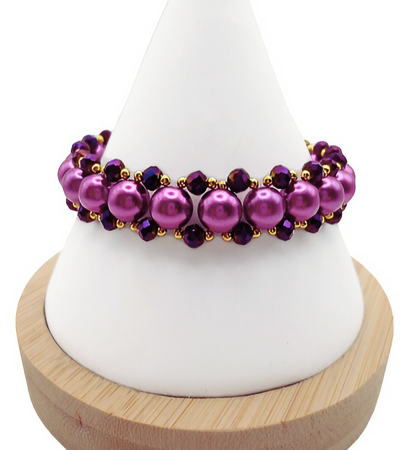 Handmade wide bracelet featuring large lustrous purple pearls and deep metallic purple faceted crystals woven together with gold seed bead accents in a full rounded cluster design, displayed on a white cone bracelet stand with a wooden base against a white background.