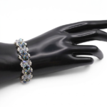 Handmade opal and silvery grey aurora borealis AB crystal and white pearl bracelet featuring colour-shifting iridescent crystals and small white pearls woven in a repeating floral cluster pattern with silver-tone bead accents, displayed on a black cone bracelet stand with a wooden base against a white background.