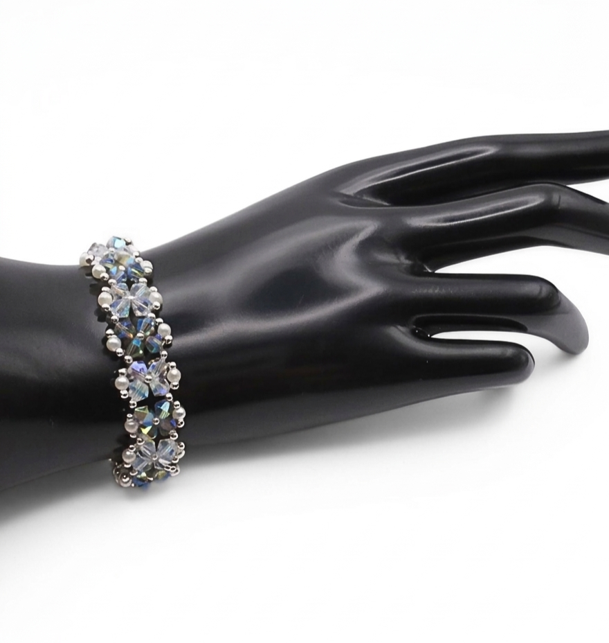 Handmade opal and silvery grey aurora borealis AB crystal and white pearl bracelet featuring colour-shifting iridescent crystals and small white pearls woven in a repeating floral cluster pattern with silver-tone bead accents, displayed on a black cone bracelet stand with a wooden base against a white background.