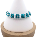 Handmade bracelet featuring teal faceted crystals arranged in a repeating four-petal flower pattern with white pearl centres and borders, and silver seed bead accents, displayed on a white cone bracelet stand with a wooden base against a white background.